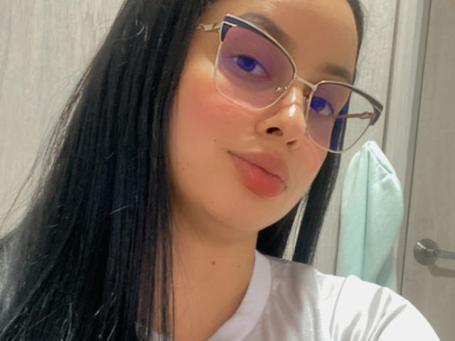 EmilyParkerr with long black hair, purple eyeshadow, and glasses, wearing a white shirt, has pinkish lips and a small nose, looking at the camera with a slight smile.