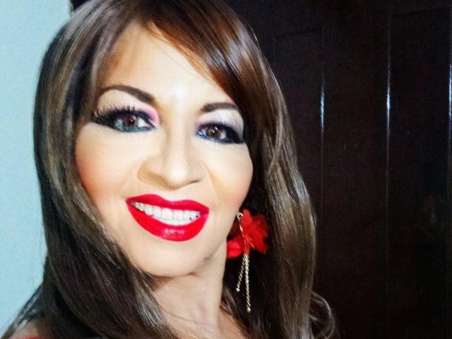 SexyLilith com cabelos castanhos brilhantes, batom vermelho, delineador preto marcante, sombra rosa e brincos de flores vermelhas, sorrindo alegremente. Seus seios estão levemente visíveis sob uma blusa preta..