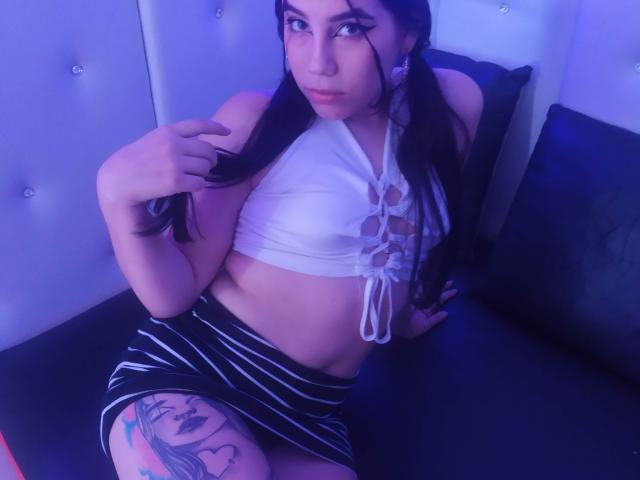 AdharaBamd con cabello negro largo, top blanco corto que muestra los pezones, falda a rayas negras y blancas, tatuaje en el muslo de un rostro de mujer, sentada en una cama negra, con iluminación azul..