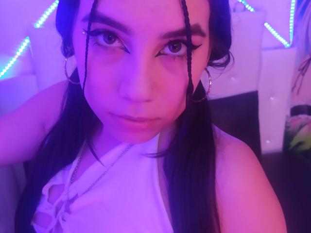 AdharaBamd con cabello negro largo, iluminación púrpura-azul, cejas gruesas, blusa blanca, aros grandes, trenzas en el rostro, mirando a la cámara, labios ligeramente fruncidos, collar, labios ligeramente entreabiertos..