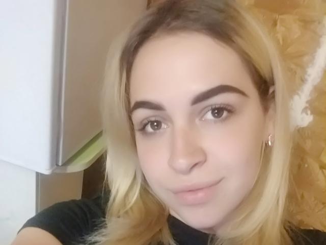 GinaLettie, con capelli biondi, sopracciglia scure, pelle chiara, naso piccolo, sorriso discreto, indossa una maglietta nera, piccoli orecchini dorati, occhi marroni, trucco leggero, guarda la fotocamera..