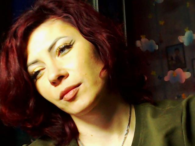 JuliaIce, con cabello rojo ondulado, delineador de ojos amarillo y la lengua ligeramente afuera, vestida con una camisa verde y un collar, luciendo juguetona. Sus ojos están medio cerrados..