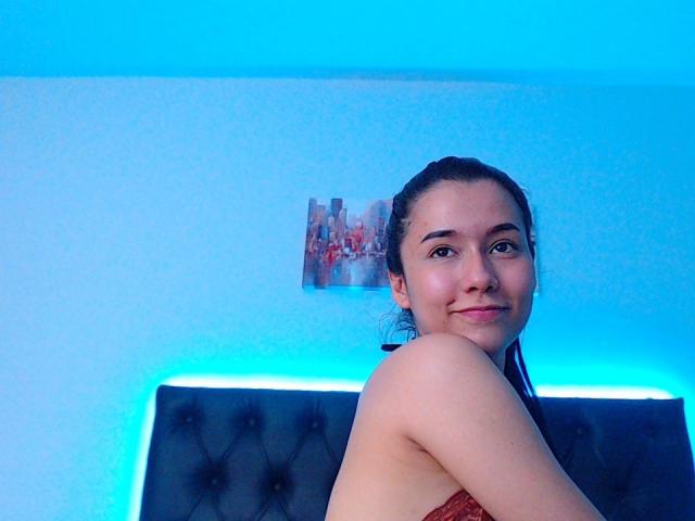 DeisyWest, avec ses cheveux noirs attachés en queue de cheval, souriante, montrant son épaule nue et son soutien-gorge rouge, est assise devant une tête de lit éclairée en bleu. Ses fesses sont partiellement visibles..