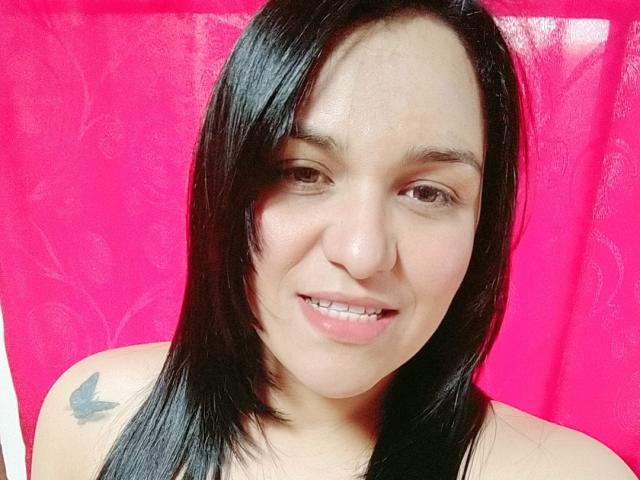 VivianaThompson has black hair, light skin, brown eyes, small nose, smiling with slightly open mouth, showing teeth, and a small bird tattoo on her right shoulder.