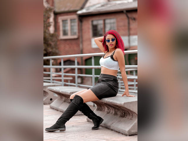 VioletaSteel with red hair, wearing sunglasses, white sports bra, black skirt, and knee-high black boots, sitting on a concrete bench, hand in hair, booted legs crossed.