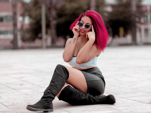 VioletaSteel, con cabello rosa brillante, gafas de sol azules, un top blanco, una falda negra y botas negras hasta la rodilla, sentada en un suelo de baldosas, con las manos cerca de la cara, y un tatuaje en el brazo izquierdo..
