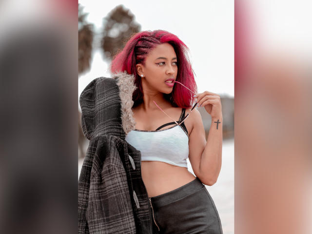 VioletaSteel mit rosa Haaren, zu Zöpfen geflochten, lutscht an einem Lolli und trägt einen weißen Sport-BH, eine schwarze Jacke und schwarze Hosen. Sie hat ein Kreuz-Tattoo auf ihrem rechten Arm..