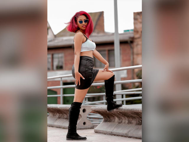 VioletaSteel with red hair, blue eyes, wearing a white crop top, black leather skirt, and knee-high black boots, standing with one leg up, showing her ass.