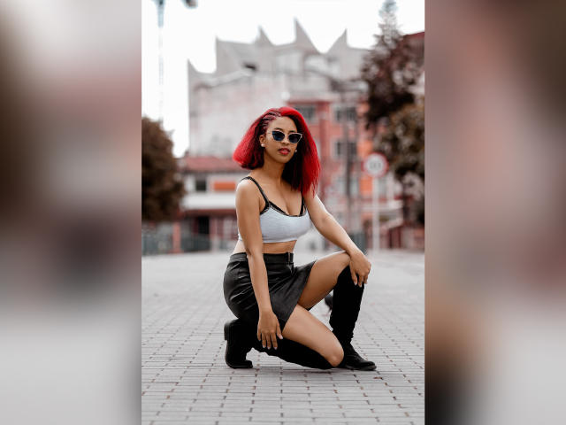 VioletaSteel met rood haar, een zonnebril, een witte crop top, een zwarte rok en kniehoge laarzen, hurkt op een straat in de stad en laat haar borsten en billen zien..