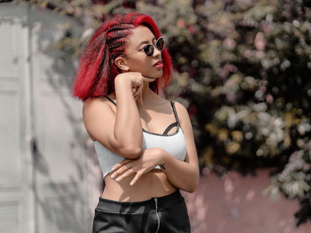 VioletaSteel, com cabelo trançado vermelho, usando óculos de sol, um top curto cinza que mostra seus seios, calças de couro pretas e um zíper, em pé com os braços cruzados, olhando para o lado..