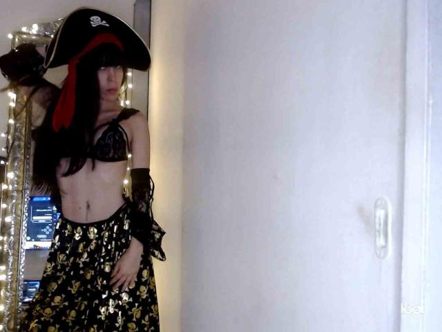 SeleneBrigth, con cabello negro largo, un sombrero de pirata, un sujetador de encaje negro y una falda con estampado dorado, mostrando sus pequeños senos y su abdomen plano, está de pie en una habitación con poca luz..