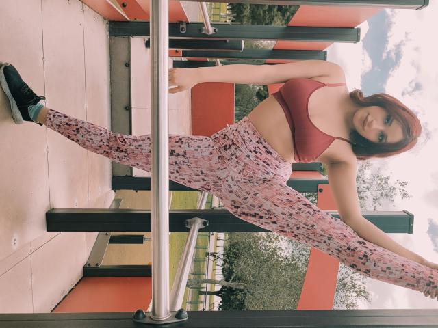 EvangelineBlossom has red hair, a red sports bra, and pink patterned leggings, doing a wide split on a metal pole, wearing black sneakers, in an outdoor gym.