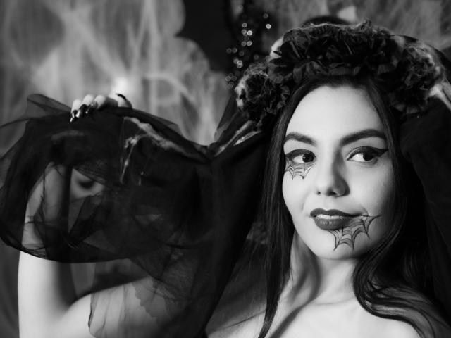 Photo en noir et blanc d'AshlyChloe avec des tatouages en forme de toile d'araignée sur ses joues, un maquillage foncé, de longs cheveux, portant un voile noir transparent et une couronne de fleurs, avec une expression satisfaite..
