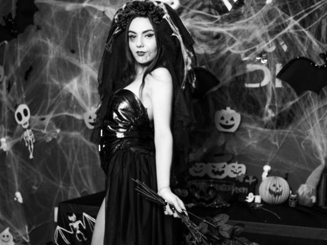 AshlyChloe mit dunklem Make-up, trägt ein glänzendes, metallisches, trägerloses Oberteil, ein schwarzes Kleid und eine Spinnennetz-Haarspange, hält einen Fledermaus und befindet sich in einem gruseligen Halloween-Ambiente mit Kürbissen..