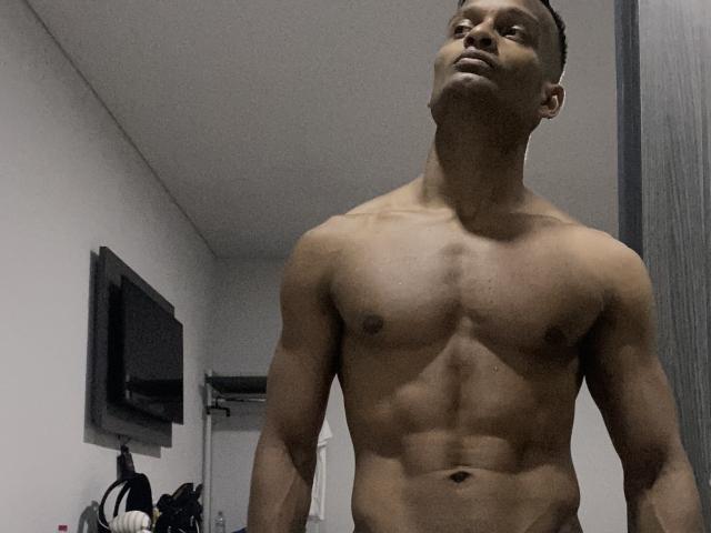 JacksonCooper, shirtless, muscular, dark-skinned, standing with a slight upward gaze, defined abs, short curly hair, bare chest, right arm down, left arm slightly raised.