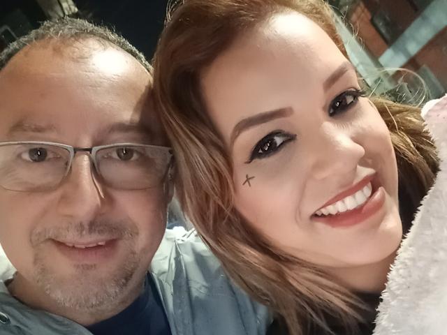AlexKaren heeft bruin haar, zwarte eyeliner en een kleine kruistatoeage onder haar rechteroog. Ze glimlacht met witte tanden, en een dikke man met een bril staat naast haar..