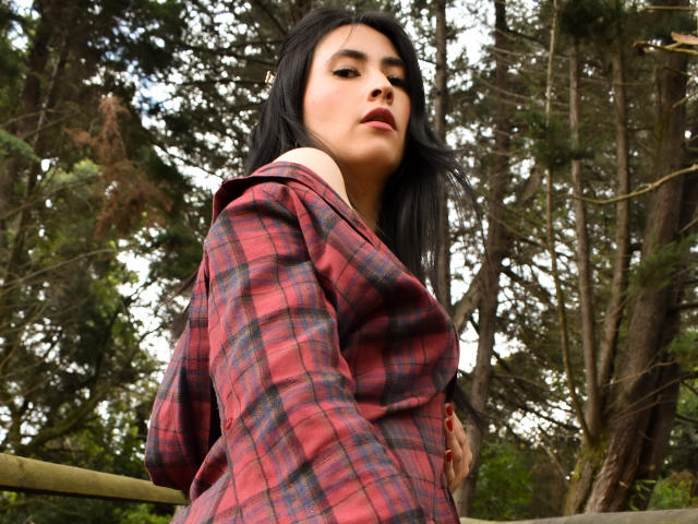 JainaRogers with long black hair, wearing a red plaid shirt, looking down, hands on her hip, standing in a forest, her face partially shadowed by trees.