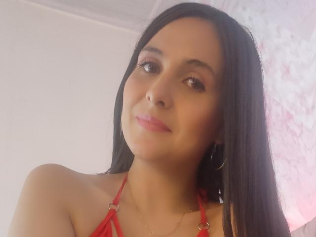 Ambeer, capelli neri lunghi, top rosso a fascia, orecchini a cerchio, leggero sorriso, rossetto rosa e un viso carino, che guarda verso il basso la fotocamera. Il suo seno è piccolo e carino..