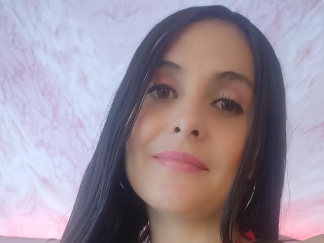 AmbeerBlack ha lunghi capelli neri, rossetto rosa e orecchini a cerchio, sorride con le labbra rosa leggermente dischiuse, e le sue ciglia sono visibili su uno sfondo rosa e bianco tenue..