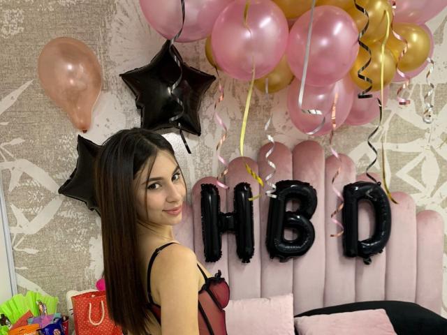 NikoleFoster with long brown hair, wearing a pink and black sheer lingerie top, looking over her shoulder with a slight smile, surrounded by pink and gold balloons.