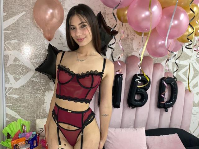 NikoleFoster with long brown hair, wearing a red and black lace lingerie set, standing in a room with pink and gold balloons, smiling at the camera. Boobs visible.