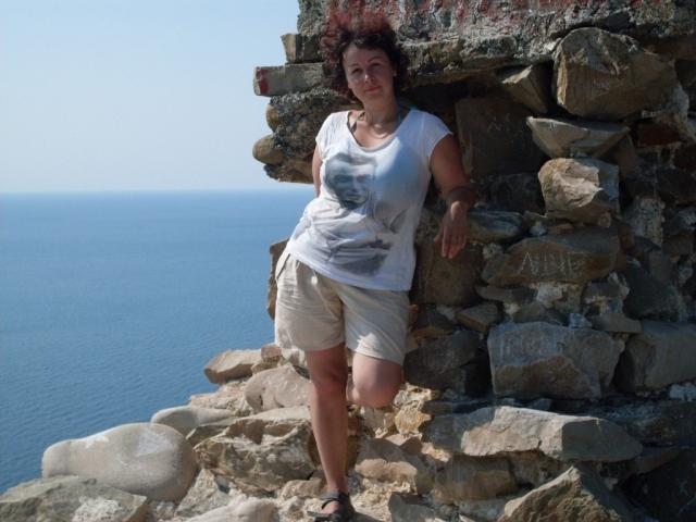 ErinSmart a des fesses rebondies, elle se tient contre un mur de pierre, vêtue d'une chemise blanche mouillée et de shorts beige, et regarde l'appareil photo avec une jambe levée..