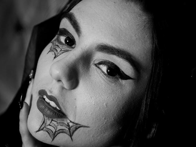 AshlyChloe's face in black-and-white, showing spiderweb tattoos on her nose and lower lip, with bold eyeliner, dark lipstick, and her hand near her chin, looking intense.