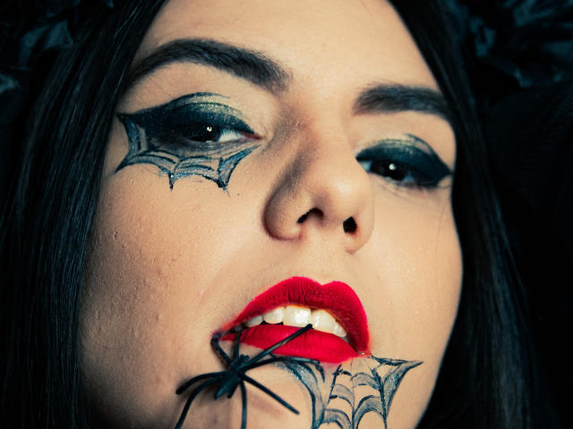 O rosto de AshlyChloe, com maquiagem escura, tatuagens de teias de aranha nas bochechas e batom vermelho. Ela está mordendo uma aranha preta enquanto olha para cima com olhos verdes e sobrancelhas pretas..