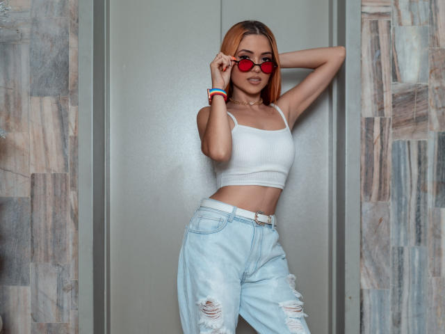 Maisy Warner, avec ses cheveux orange, portant des lunettes de soleil rouges, un crop top blanc et un jean déchiré, appuyée contre un mur, les bras levés, montrant sa petite poitrine et ses hanches fines..