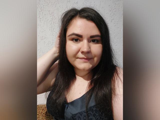 BeckyAndEllen, avec de longs cheveux noirs, des yeux marron et un piercing au nez, portant un haut en dentelle noire, une main dans les cheveux, souriant légèrement, mur blanc texturé en arrière-plan..