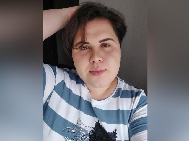 BeckyAndEllen, met kort bruin haar, blauw-groene ogen en een lichte glimlach, draagt een wit en blauw gestreept shirt en heeft één arm achter haar hoofd..