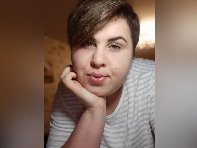 BeckyAndEllen, avec des cheveux bruns courts, portant une chemise blanche rayée, appuyant son menton sur sa main, souriant doucement à la caméra. Ses yeux verts sont légèrement plissés..
