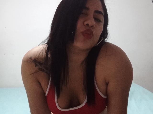 Anaii, avec ses cheveux foncés, son débardeur rouge et sa poitrine dégagée, faisant la moue avec ses lèvres rouges, a un tatouage sur son épaule droite, porte des boucles d'oreilles pendantes, sur un fond blanc uni..