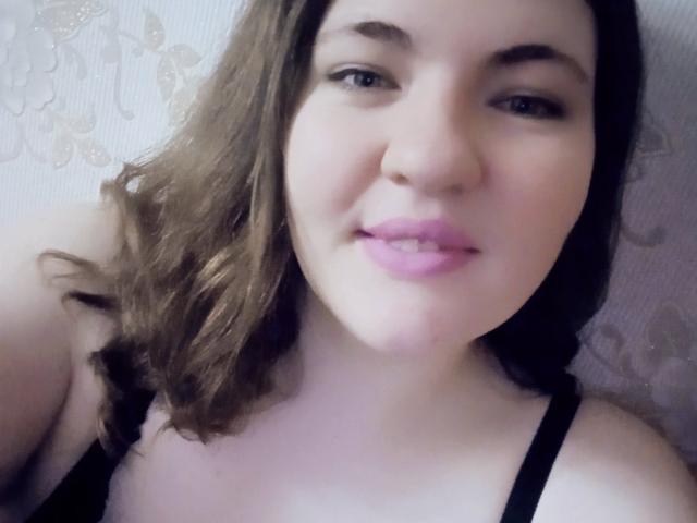 AglayaFame com batom rosa, cabelo castanho ondulado e sutiã preto, sorrindo para a câmera, mostrando seus seios e rosto levemente rechonchudo. Pele macia e clara, olhos azuis..
