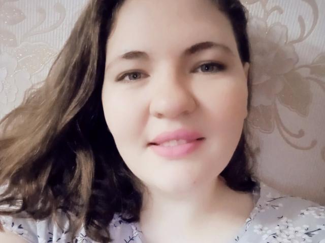 AglayaFame a les lèvres roses, les yeux marron et les cheveux bruns ondulés, et elle porte une chemise blanche à motifs floraux. Son visage est légèrement rougi, et elle regarde l'appareil photo avec un sourire discret..