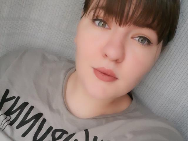 AglayaFame mit kurzen, braunen Haaren, grünen Augen und rosafarbenem Lippenstift, trägt ein graues Hemd und blickt mit einem leichten, neutralen Ausdruck in die Kamera..