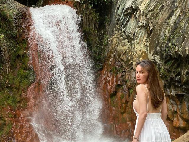 BeautyQueenTs with long brown hair, white dress, standing right, looking back at waterfall. Water cascades down rocky, moss-covered cliff. She has small tits, cute face, and a sexy look.