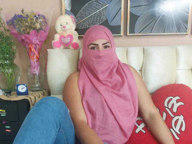 Sahily, una donna formosa con la pelle di colore marrone medio, indossa un hijab rosa e un asciugamano rosa che le copre il viso e il petto, è seduta su un divano bianco con jeans blu..