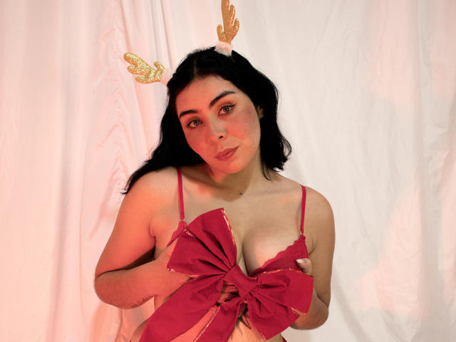 DannaDee met donker haar, gekleed in rode lingerie en een gouden hoofdband met gewei, houdt een grote rode strik boven haar borsten, en staat voor een witte gordijn..