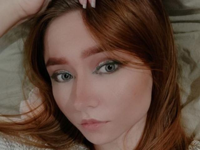 AnnaTavia has red hair, green eyes, and light makeup. She's lying down with her hand in her hair, looking serious. Her tits are partially visible. Her ass is not.