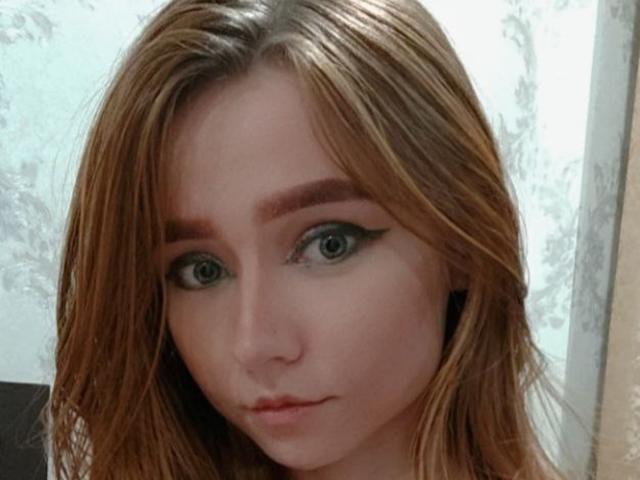 AnnaTavia has pale skin, green eyes with thick black eyeliner, and long brown hair. Her eyebrows are dark and slightly raised, and she has a neutral expression.