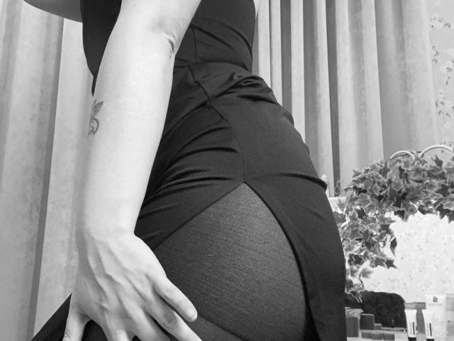 KarKutie's hand rests on her ass, wearing a tight, black dress with a sheer panel. She has a small tattoo on her upper arm. Black-and-white photo.