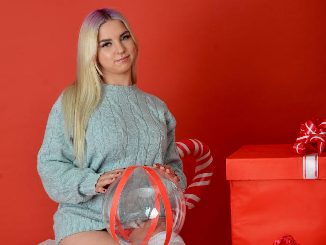 KimDenilo mit blonden Haaren, einem hellblauen Pullover, hält einen durchsichtigen Ball mit roter Schleife und sitzt neben einer roten Geschenkbox mit weißer Schleife, lächelnd. Die Brüste sind unter dem Pullover sichtbar..