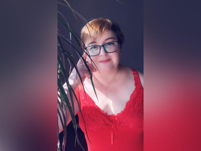 LimonkaFire, avec ses courts cheveux bruns, ses lunettes et son haut en dentelle rouge, sourit légèrement, debout entre des murs rouges et gris, avec une plante verte sur la gauche..