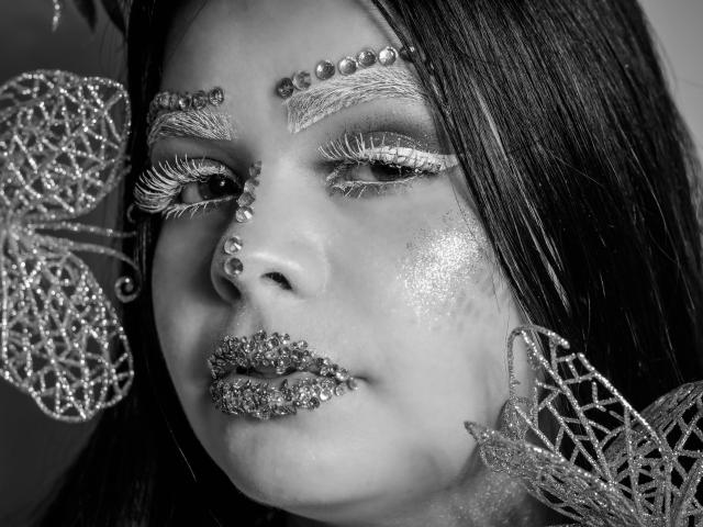 Il viso di BrithneyLee è decorato con glitter, perle sulla fronte e sul naso, e un trucco scintillante. Ha le labbra ricoperte di glitter e grandi orecchini a forma di farfalla scintillanti. Foto in bianco e nero..