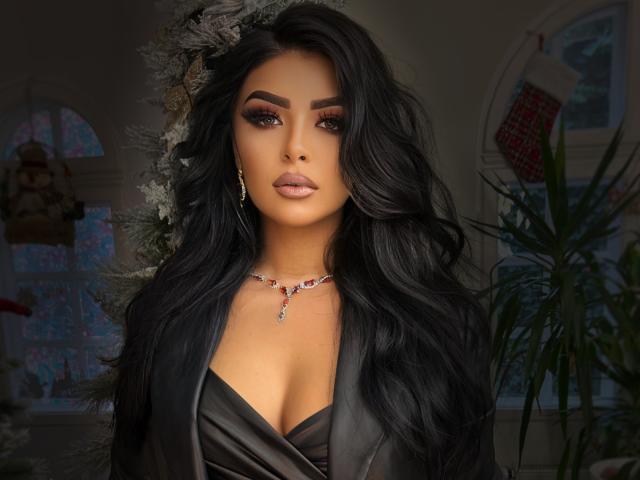 Devasha has dark, wavy hair, full eyebrows, and is wearing a low-cut black satin top showing her tits. She has a red gem necklace and is looking at the camera.