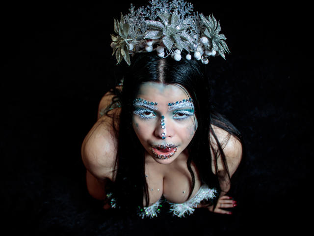 BrithneyLee, à quatre pattes, avec un maquillage bleu glacial, des paillettes sur les seins et un serre-tête en forme de flocon de neige argenté, regarde vers le haut, la bouche ouverte. Fond noir, avec un accent sur le visage et la poitrine..