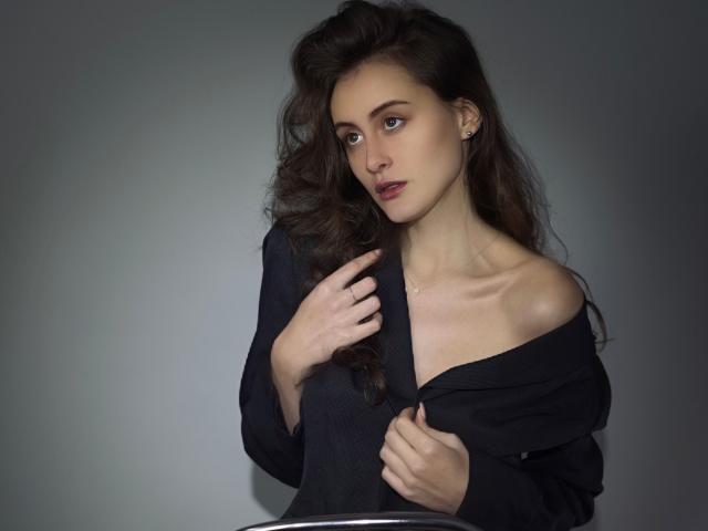 AlesyaDrons with wavy brown hair, pale skin, and small tits, wearing a black off-shoulder top, holding it with one hand, looking slightly to the side, in a photo.