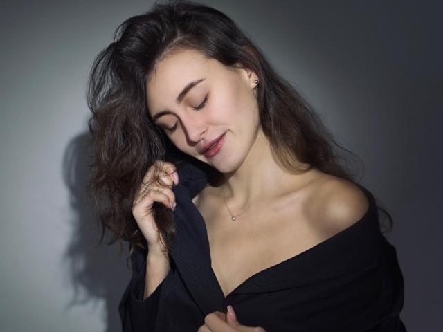 AlesyaDrons with wavy brown hair, eyes closed, smiling slightly, holding a black shirt off her bare shoulders, showing her tits, against a dark background.