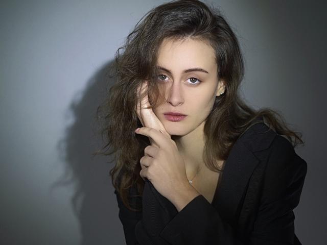 AlesyaDrons has wavy brown hair, wearing a black blazer, with a serious expression, touching her cheek with a hand, showing a shadow on the gray wall.